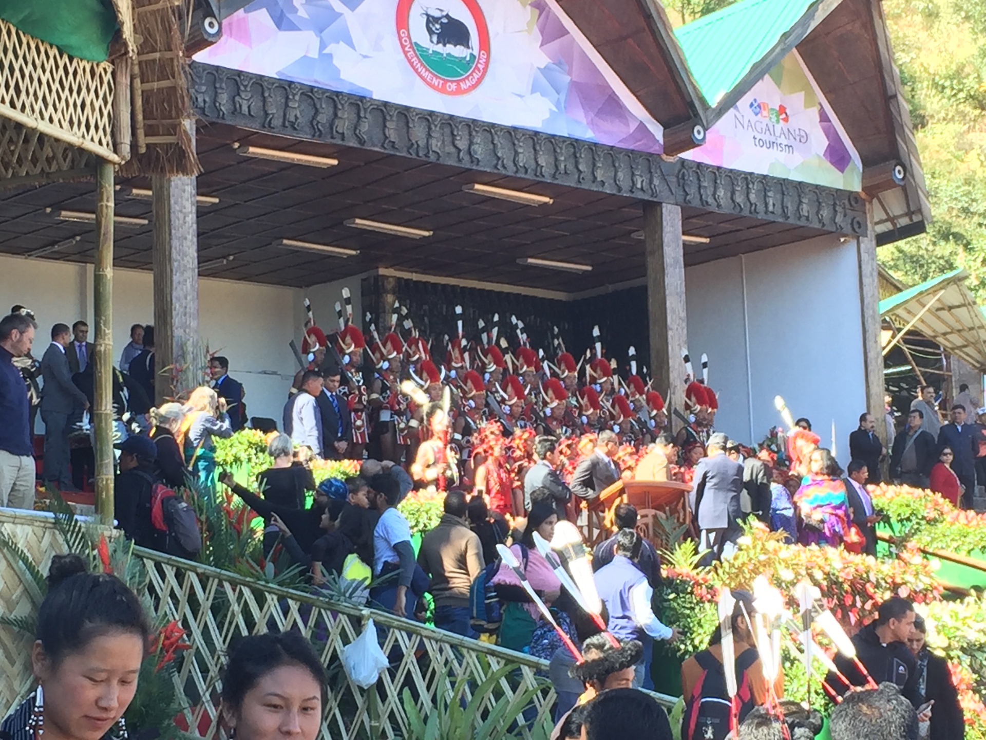 The main stage of the Hornbill Festival at Kisama Heritage Village on opening morning, with Government of Nagaland and Nagaland Tourism banners overhead and ranks of Naga guards in red-fur headdresses and tall white plumes standing behind the dignitaries