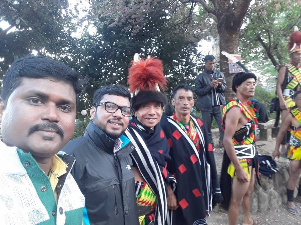 A selfie taken at the Hornbill Festival in Nagaland with the group's Indian guide beside two Naga performers in full festival attire — one wearing a red-fur headdress, the other a striped red and black shawl