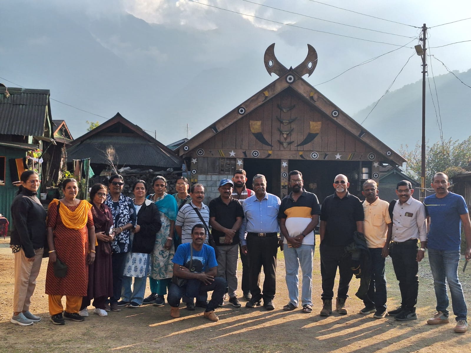 The Kerala group standing in front of a wooden Naga house in Khonoma Village, its gable carved with crossed mithun horns and traditional motifs, with the Angami hills behind