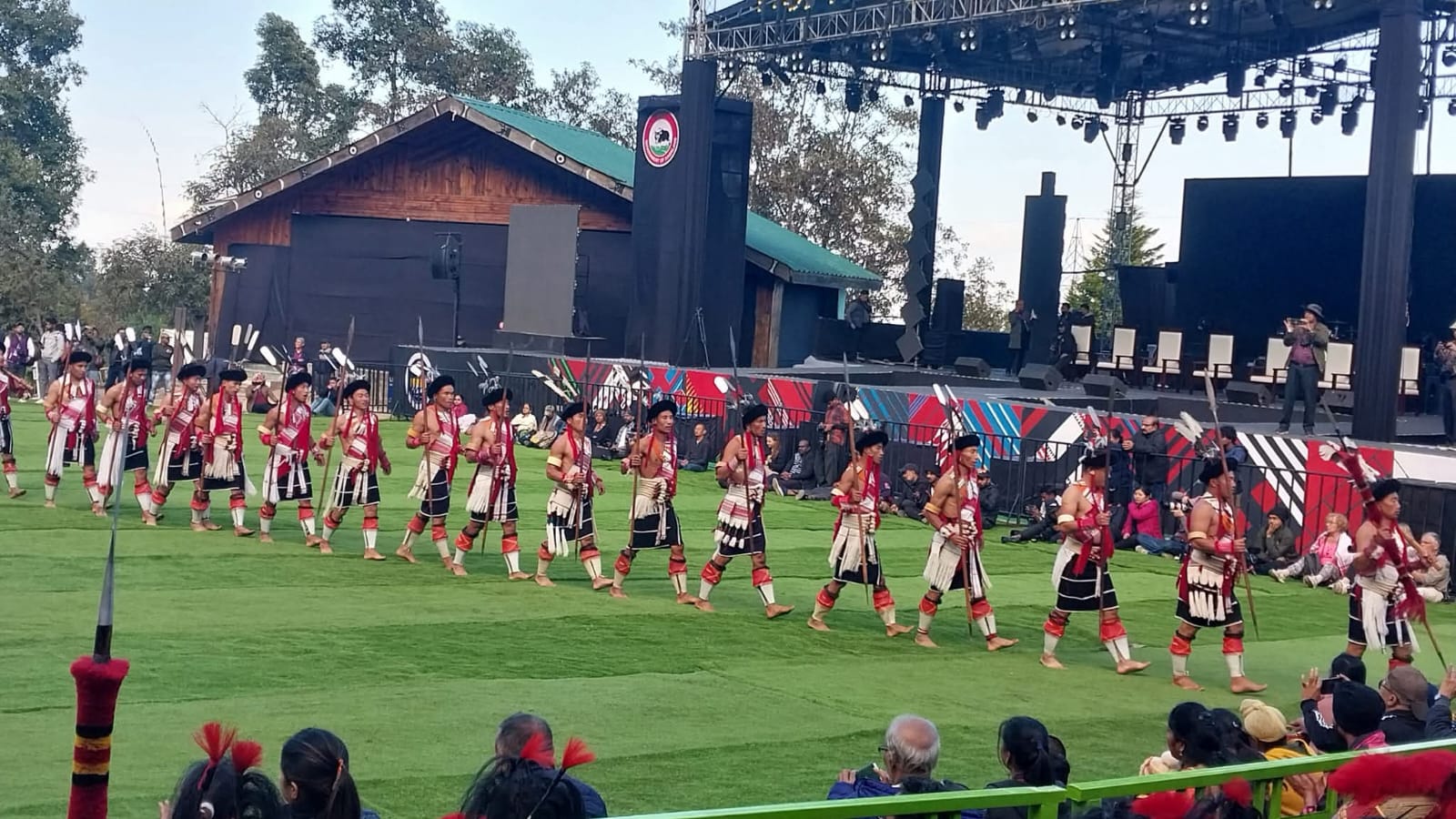 A long line of Naga warriors in bear-fur headbands, red-and-black shawls and cowrie-shell aprons marching in unison across the green in front of the main stage at the Hornbill Festival arena, Kisama