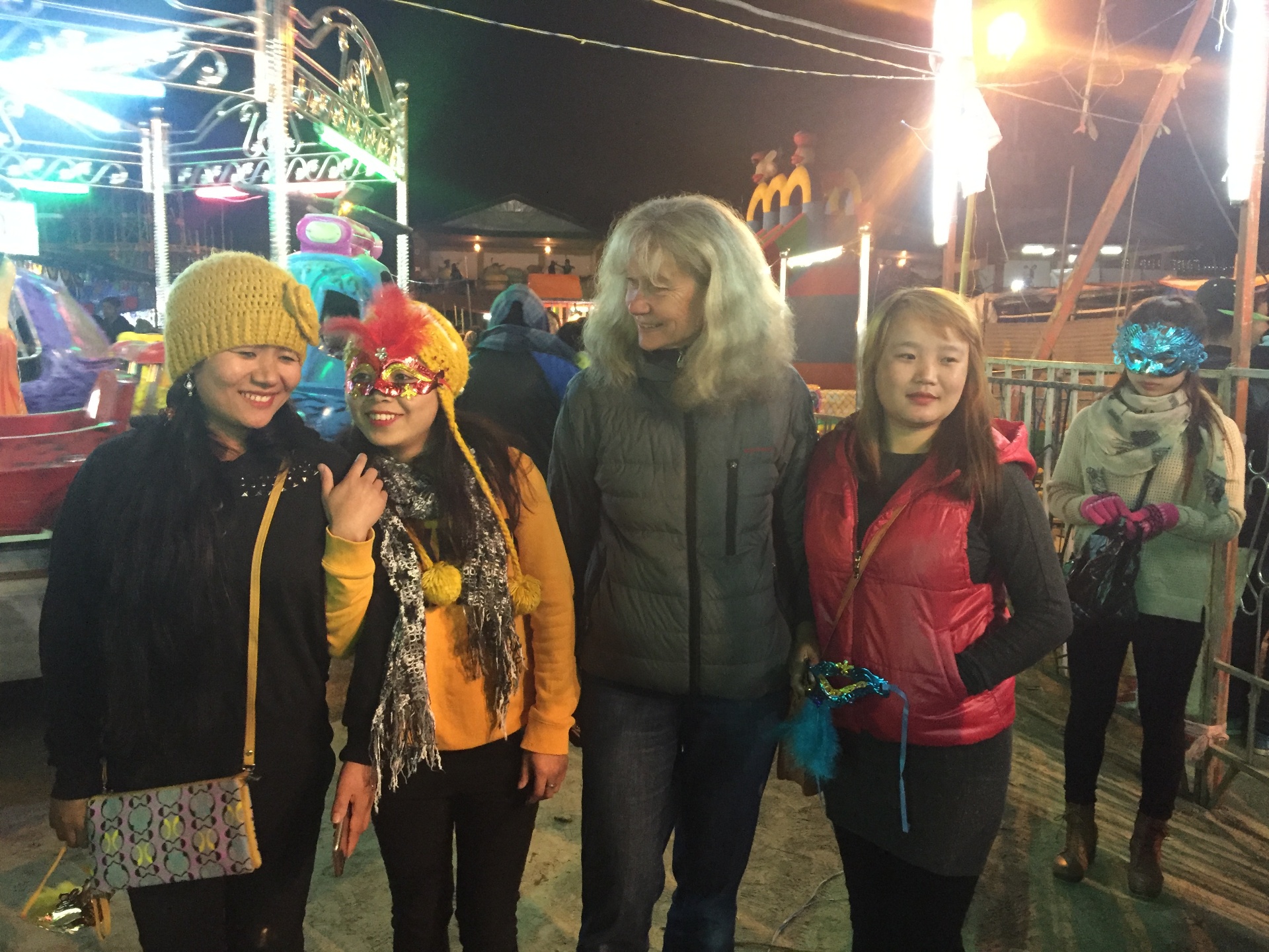 Mrs. Gulder standing with three young women from Kohima at the Hornbill Night Carnival, one of the girls wearing a red and gold sequinned Venetian-style mask, with a lit fairground ride behind them