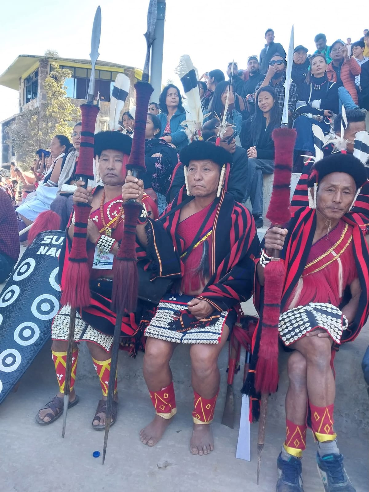 Three older Naga elders in red and black striped shawls, bear-fur headbands and beaded skirts, seated at the front of the stands at the Hornbill Festival, holding tall spears wound with red fur
