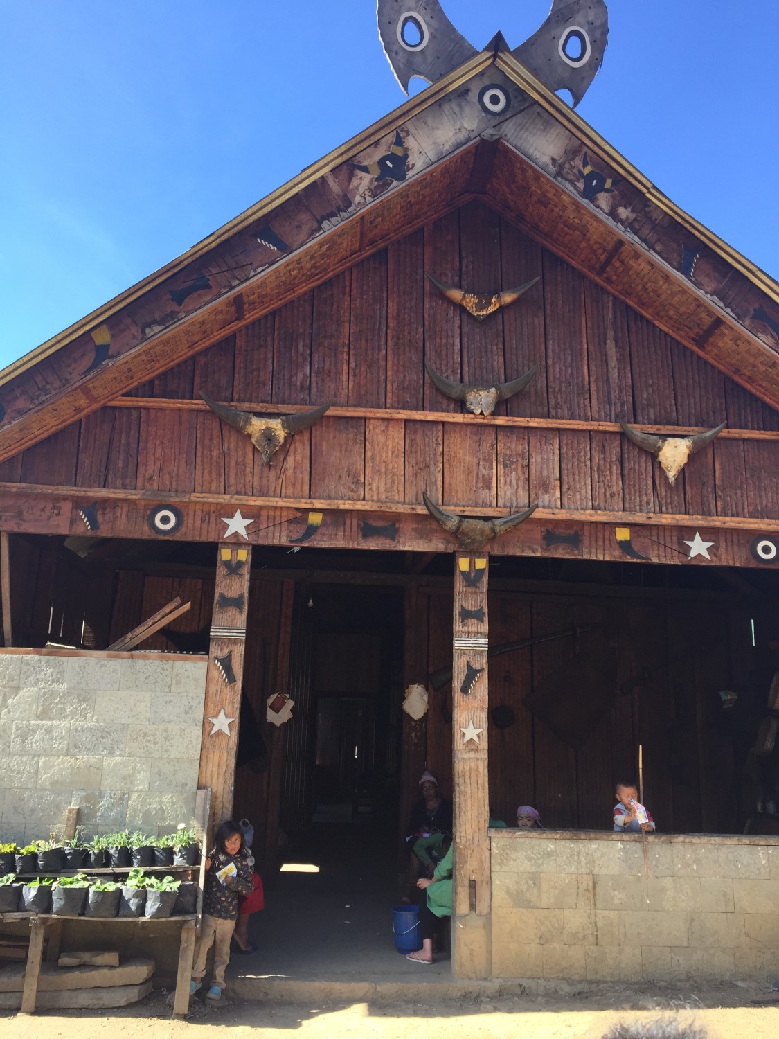 The tall carved wooden facade of a traditional Naga morung with mithun horns and painted stars along the gable and a great crown of carved wooden hornbill horns at the roof peak, with two small children at the doorway