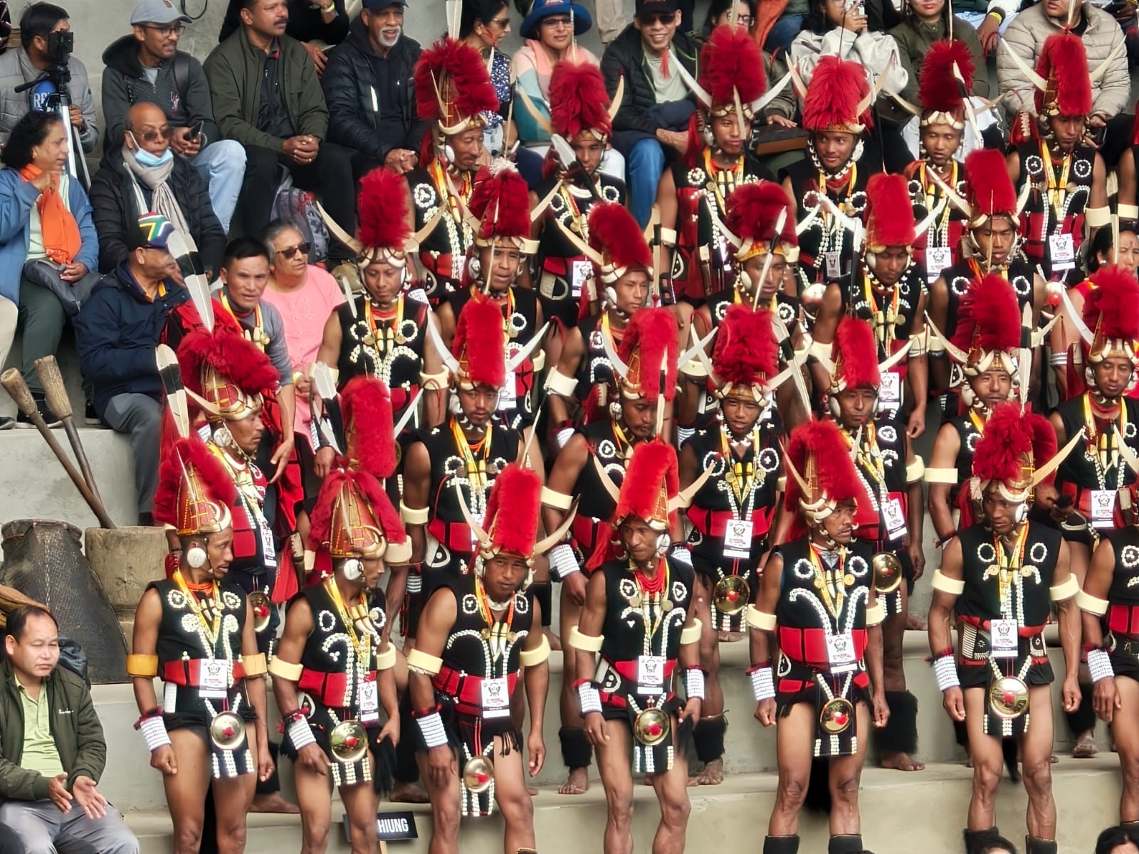 Naga warriors in red fur headdresses and mithun-horn crowns lined up in the Kisama arena at the Hornbill Festival