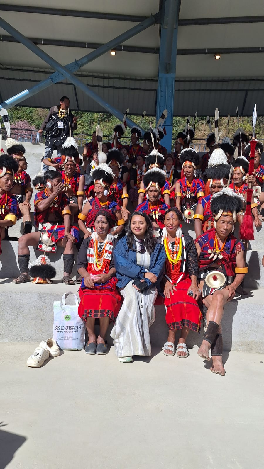 The group's local guide seated in the front row among a row of Konyak elders in traditional red shawls, boar-tusk collars and feathered headdresses on the stands at Kisama Heritage Village during the Hornbill Festival