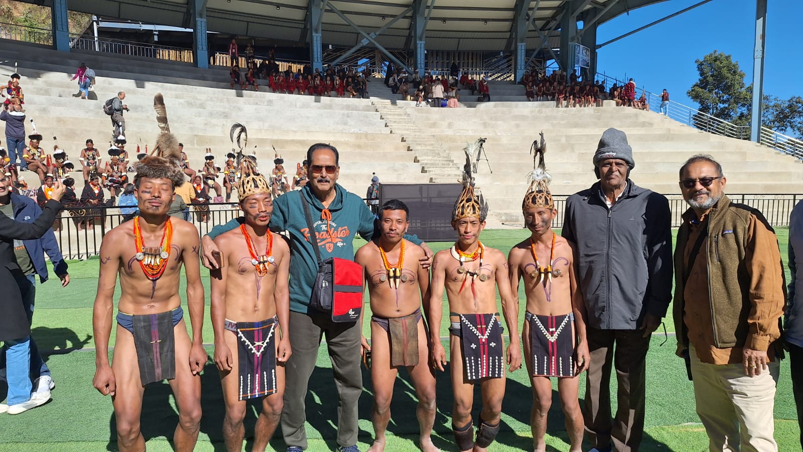 A small group of travellers from the Kerala party standing shoulder to shoulder on the Kisama arena turf with five Naga warriors in feathered wooden crown headdresses and traditional body paint, empty stone stands rising behind