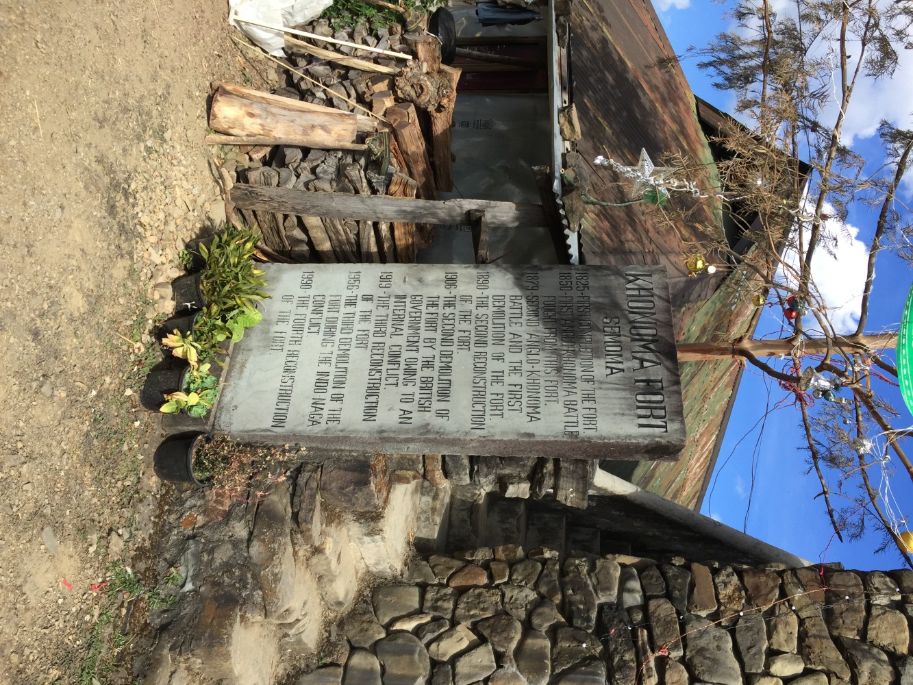 A stone memorial plaque outside the reconstructed Khonoma Fort (Semoma) in Nagaland, listing the dates of the first completion, the two Anglo-Khonoma battles, and the successive demolitions and reconstructions of the fort from 1825 to 1990