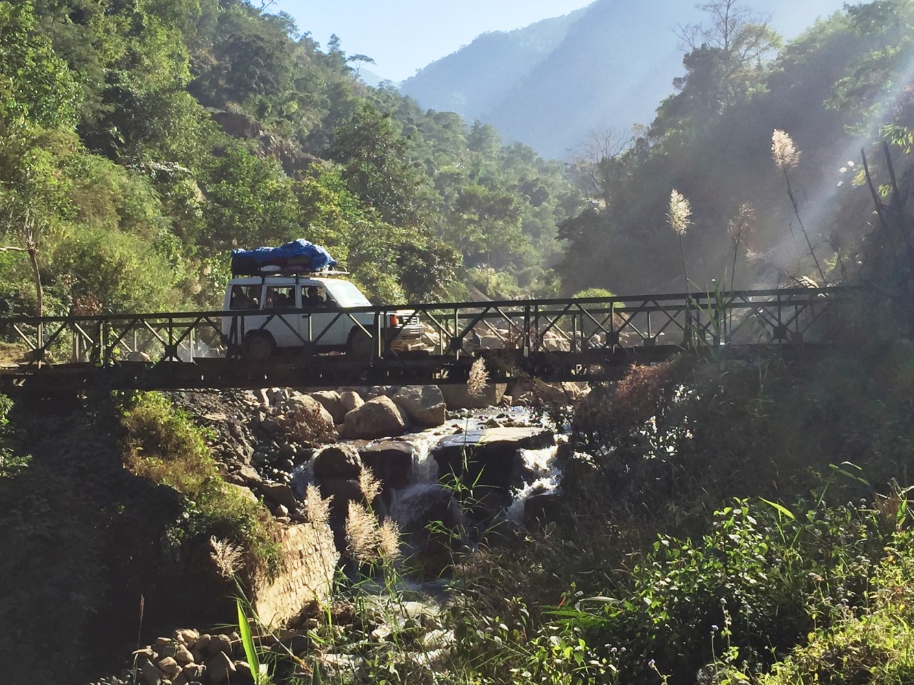 A vehicle crossing a metal Bailey bridge over a small waterfall on the mountain road between Dimapur and Kohima, with forested hills in the background