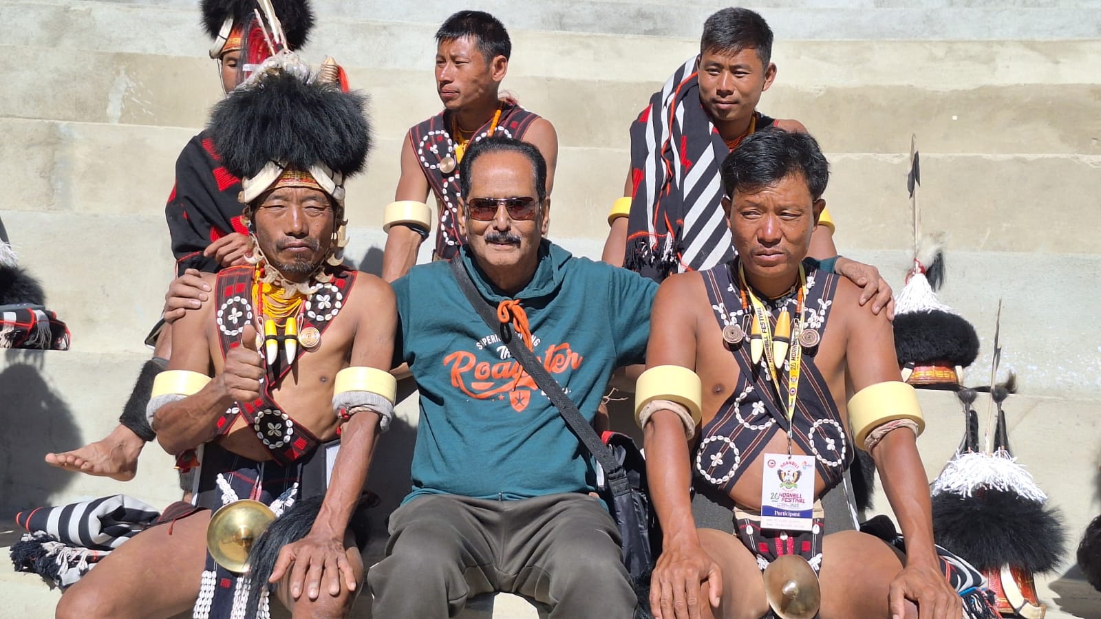 A senior traveller from the Kerala group seated between two Konyak elders in traditional dress — one wearing a tall black-fur headdress, the other in a striped shawl with heavy ivory armbands — on the stands at Kisama