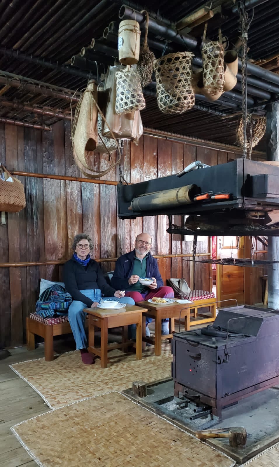 Robert and Odile eating a home-cooked lunch in a traditional wooden Mising kitchen on Majuli island, with baskets hanging from the rafters
