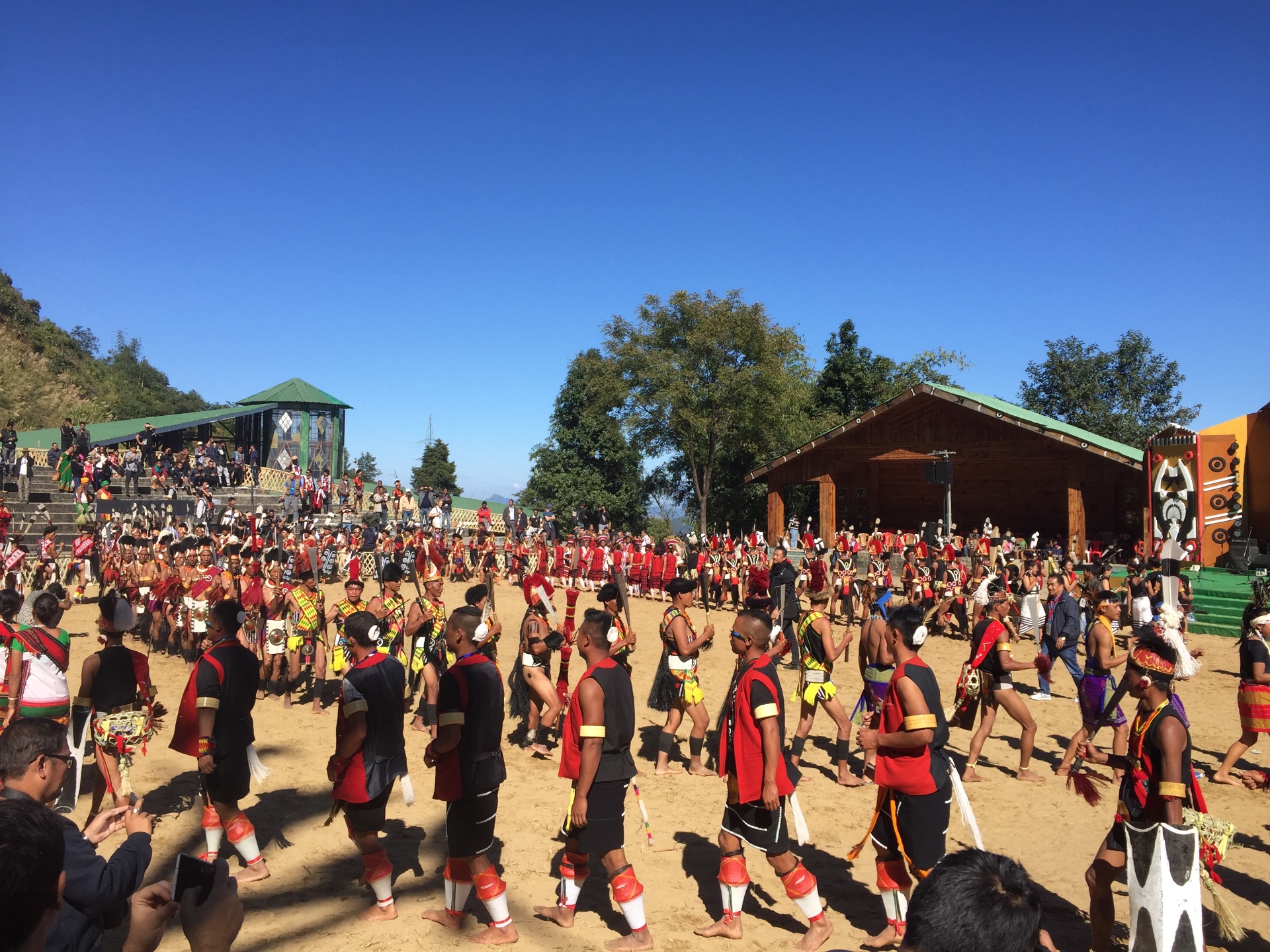 Naga warriors from several tribes in traditional attire — red and black shawls, feathered headdresses and beaded ornaments — walking across the sand arena at Kisama Heritage Village during a Hornbill Festival performance