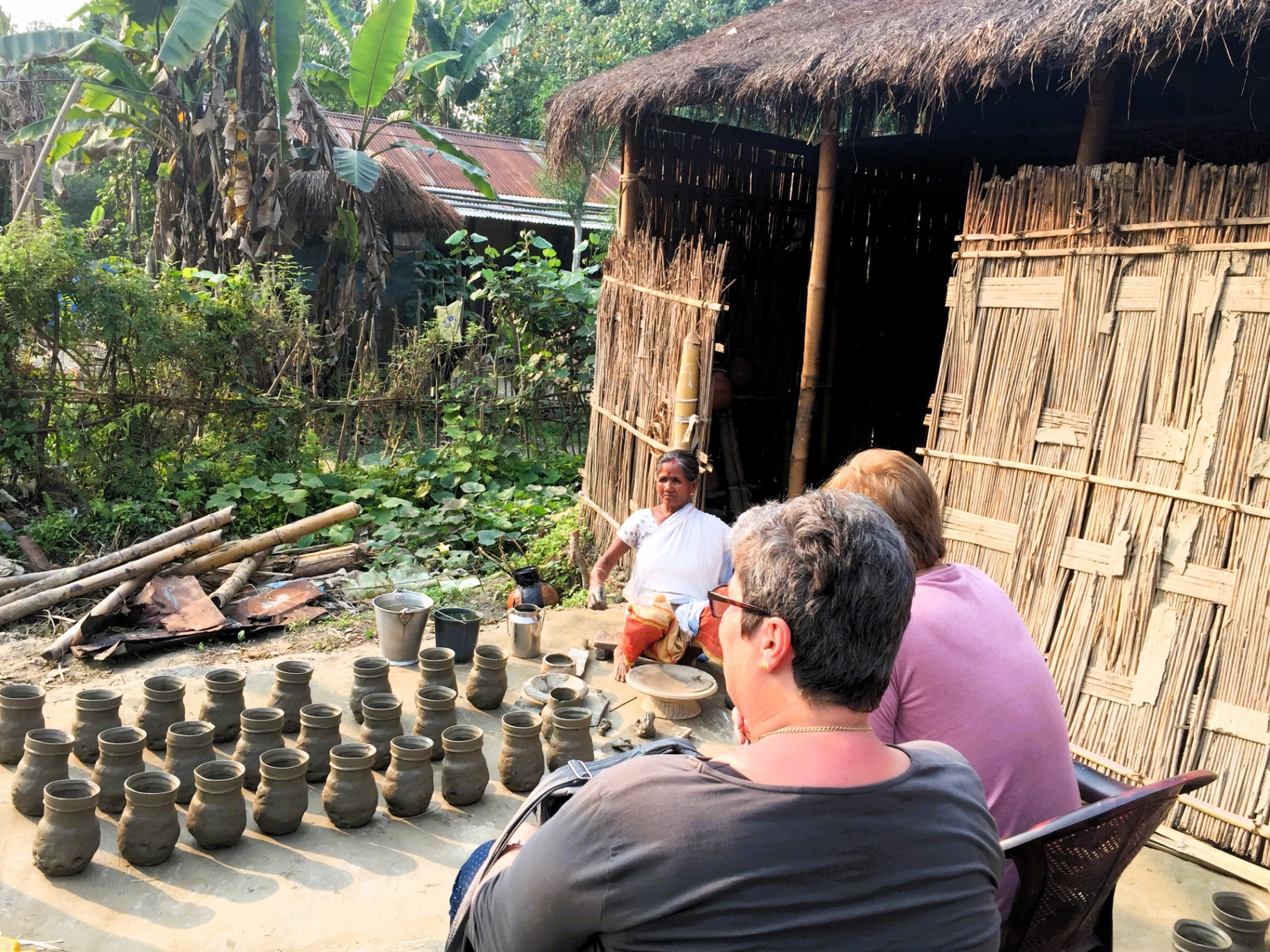 An elderly Mising potter shaping clay pots by hand outside a bamboo hut in Salmora village on Majuli island, watched by two travellers from the German group