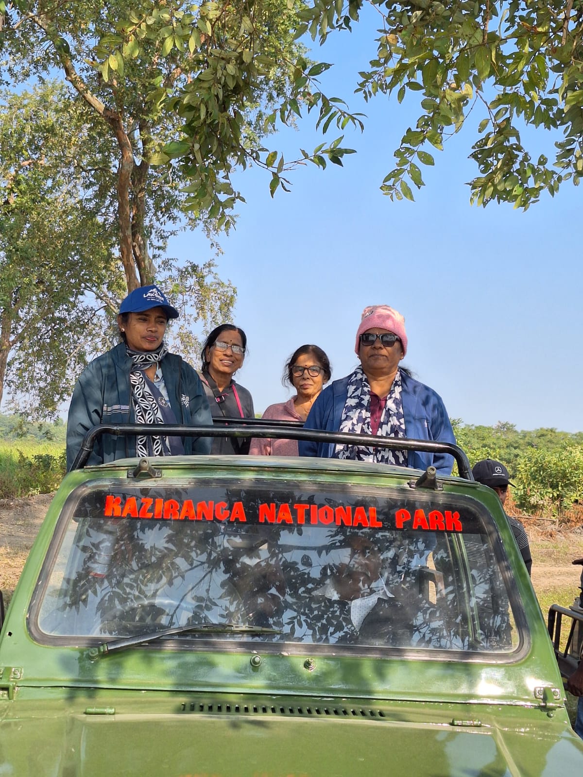 Four women from the Kerala group standing in an open Kaziranga National Park jeep during a morning safari, with the park's grasslands visible behind them