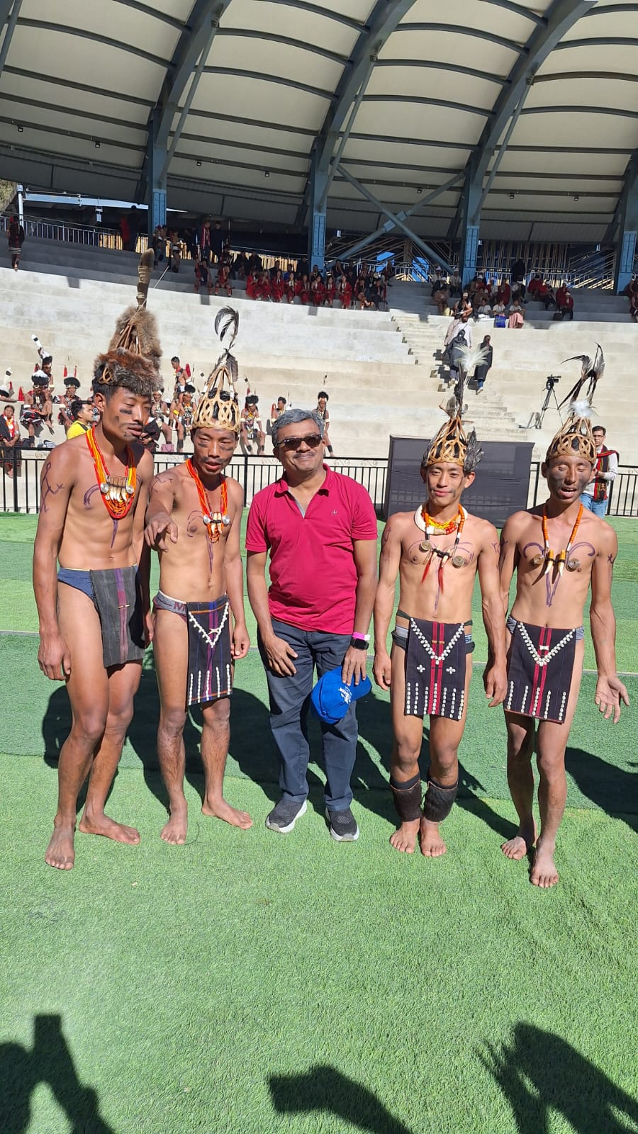 A traveller from the Kerala group in a red polo shirt standing on the Kisama arena grass with four young Chakhesang warriors in carved wooden crown headdresses and boar-tusk collars