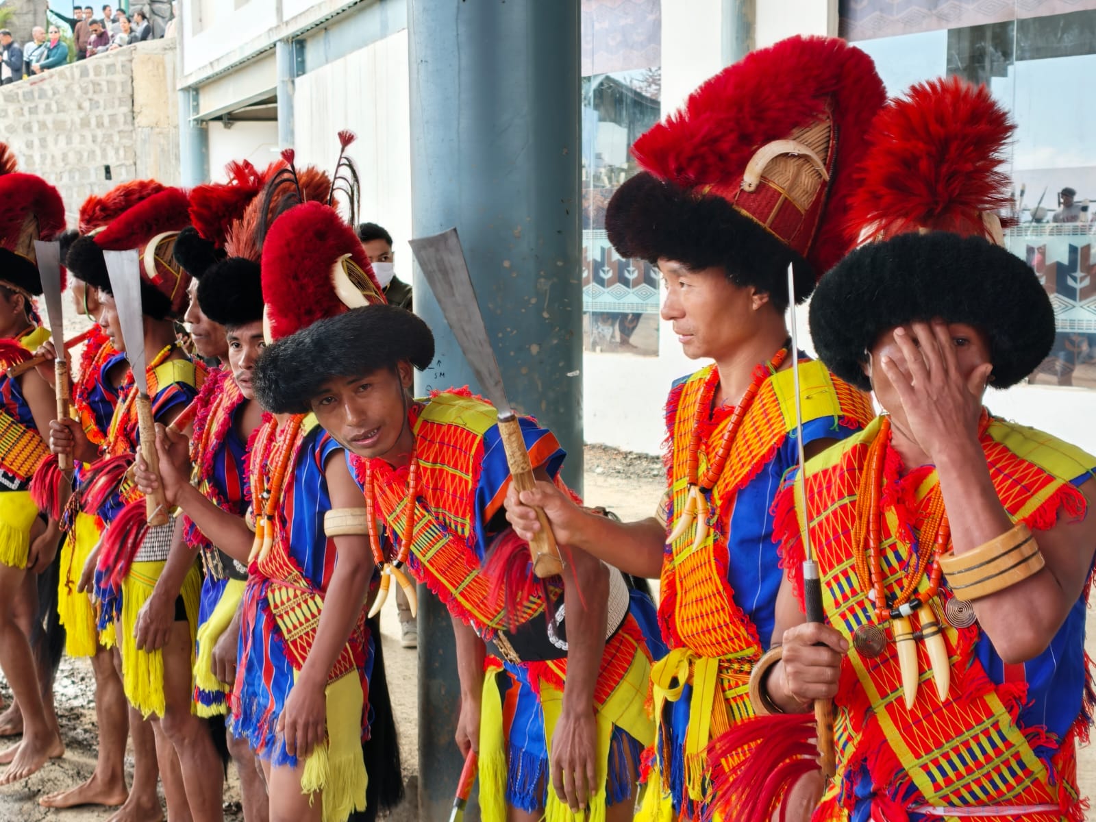 Naga performers in red plume headdresses, yellow and blue robes and ivory earrings lined up outside a morung at the Hornbill Festival