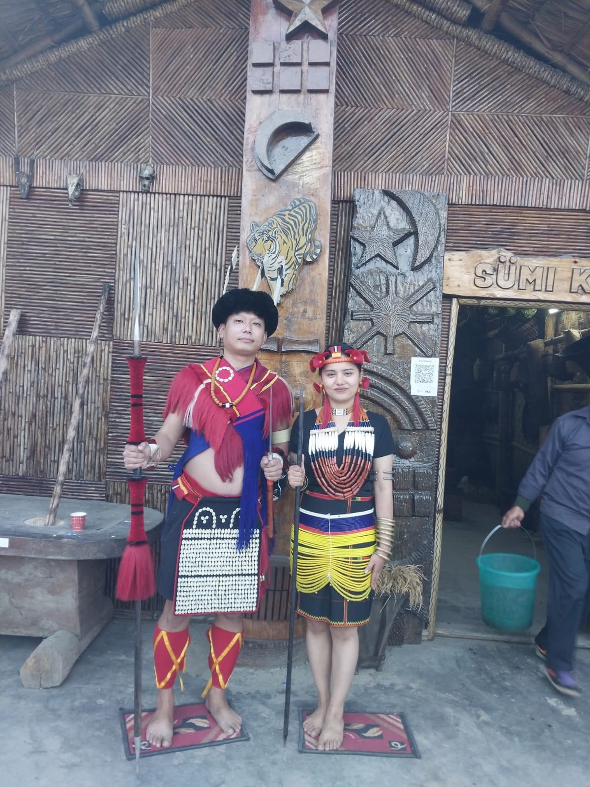 A young Sumi Naga couple in full traditional attire standing outside the Sumi Kilhomi morung at Kisama Heritage Village during the Hornbill Festival — the man with a ceremonial spear and beaded apron, the woman in a beaded skirt and shell necklace