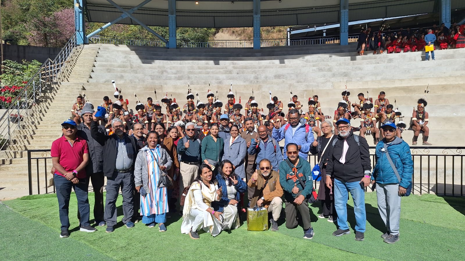 The Kerala group led by Mahim inside the Kisama arena at the Hornbill Festival, with Konyak warriors in feathered headdresses seated on the stands behind them